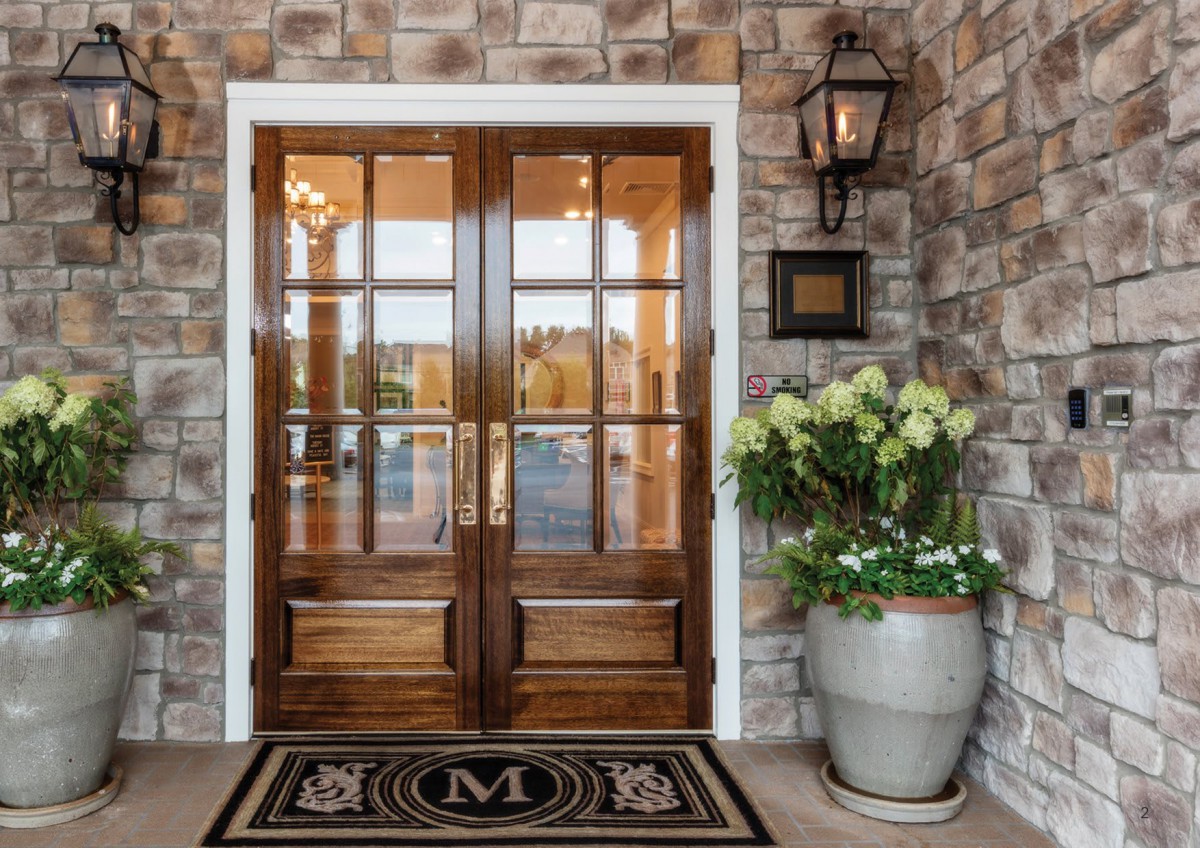 Elegant wooden double doors with a glass pane design, framed by a stone wall with a welcoming doormat and potted flowers.