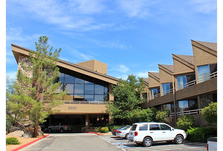 A modern office building with large windows and a parking lot in front, surrounded by trees under a clear blue sky.