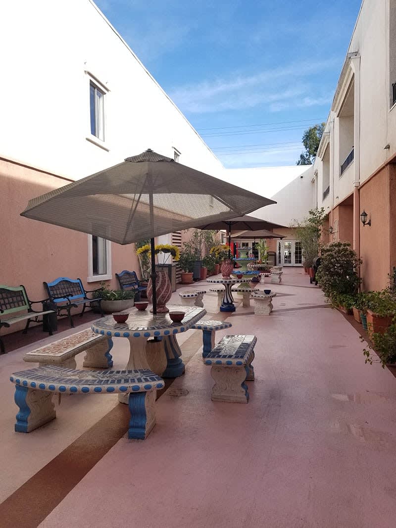 A courtyard with tables and chairs, shaded by umbrellas, surrounded by plants and buildings.