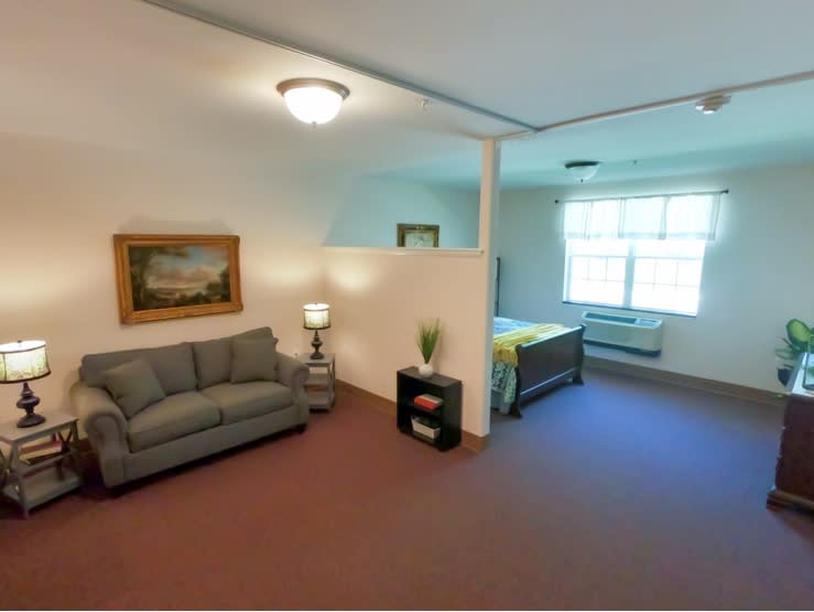 A cozy studio apartment featuring a living area with a sofa and lamp, adjacent to a bedroom space with a bed and window.