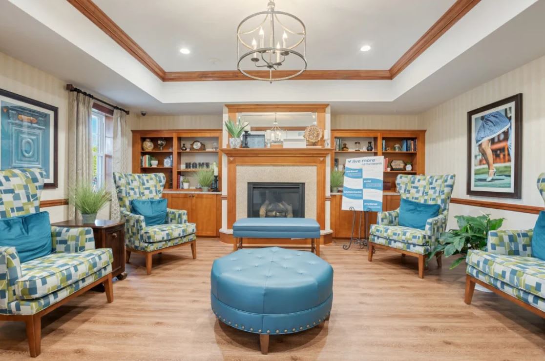 A cozy living room featuring a central fireplace, surrounded by patterned armchairs and a blue ottoman, with built-in wooden shelves adding charm to the space.