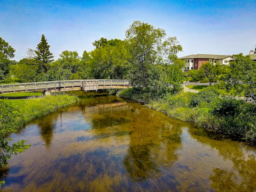 A serene landscape featuring a wooden bridge over a tranquil stream, surrounded by lush greenery and clear blue skies.