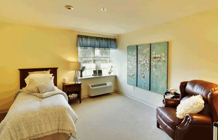 A cozy bedroom with light-colored walls, featuring a bed, nightstand, artwork on the wall, and a window with a blue valance.