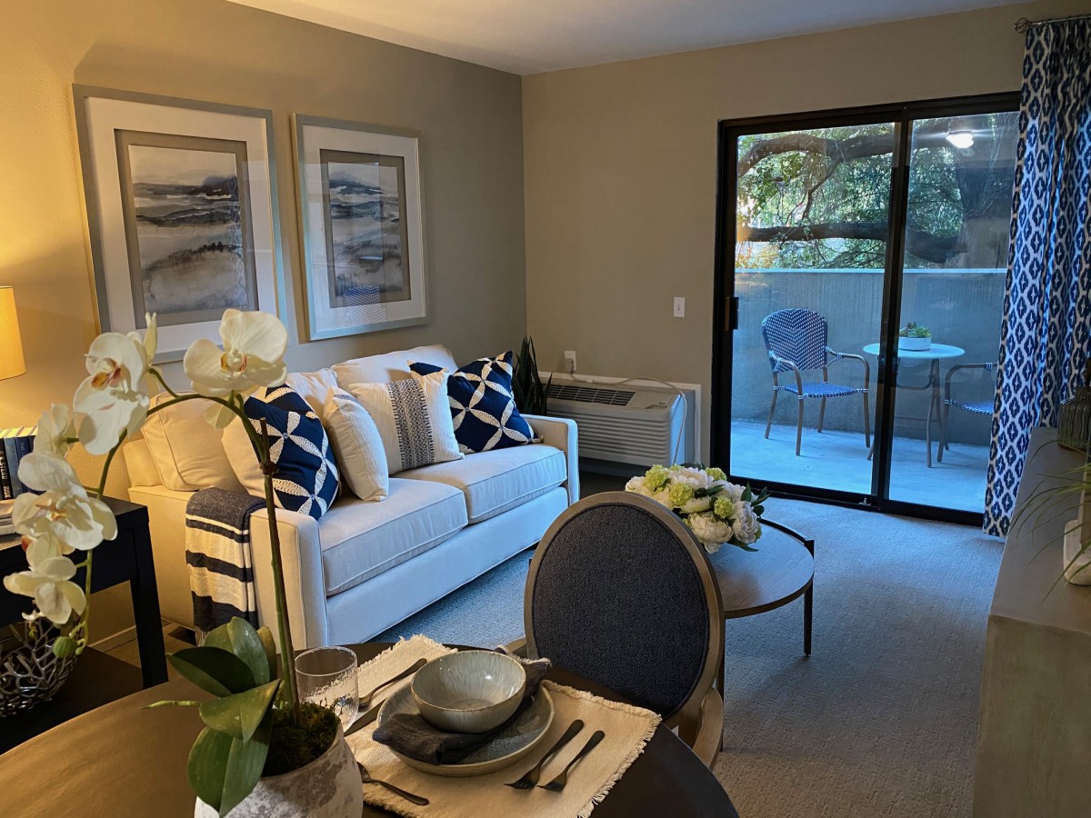 A cozy living room with a sofa, coffee table, and a dining area, leading to a balcony with outdoor seating.
