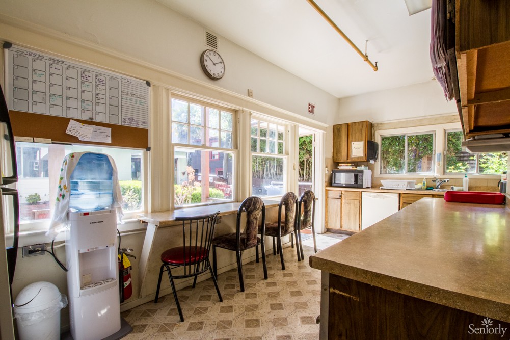 A bright kitchen with multiple chairs lined up against large windows, a countertop, and appliances including a microwave and water cooler.