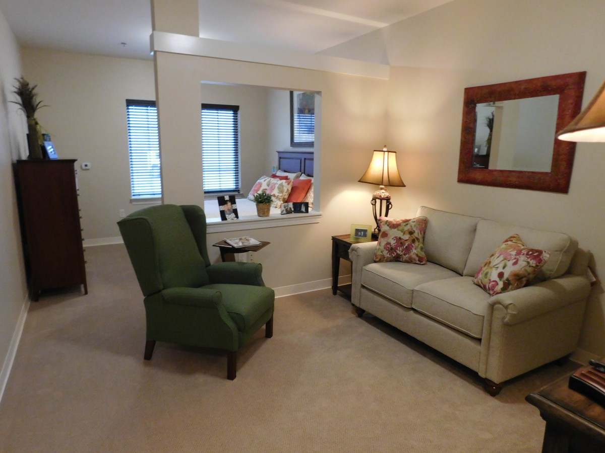 A cozy living room with a green armchair, a beige couch adorned with patterned pillows, and a warm lamp beside a mirror on the wall.
