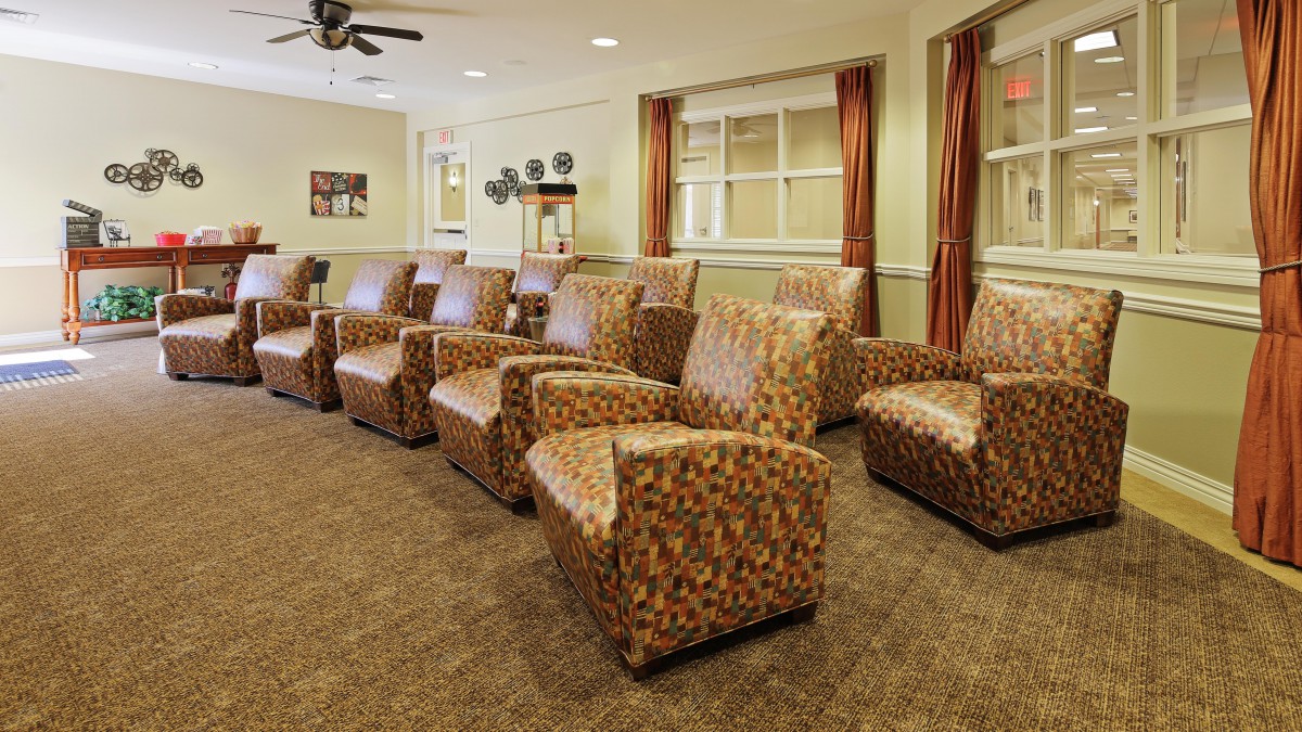 A room with multiple patterned armchairs arranged in rows on a carpeted floor, featuring large windows with curtains.