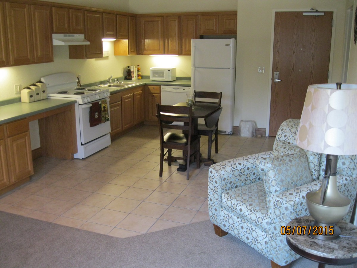 A cozy apartment kitchen with wooden cabinets, a small dining table, and an armchair.