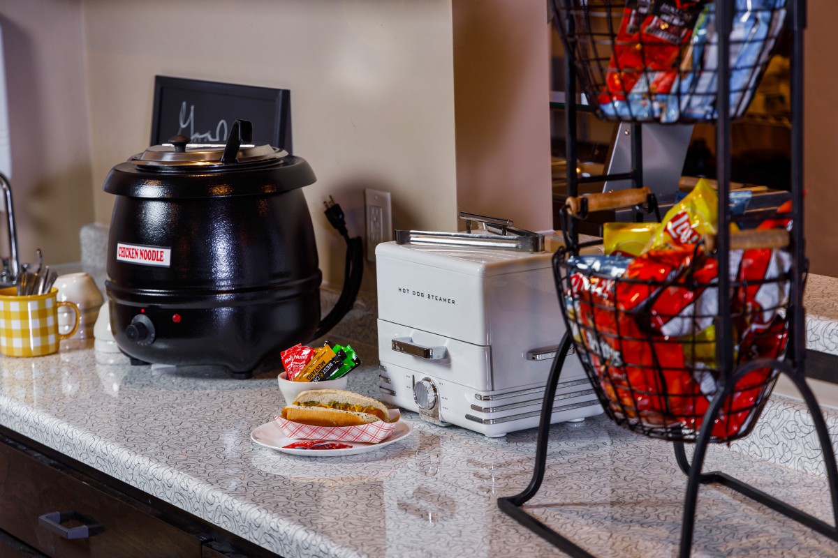 A kitchen counter featuring a soup pot, toaster, sandwich, and a rack full of chips.