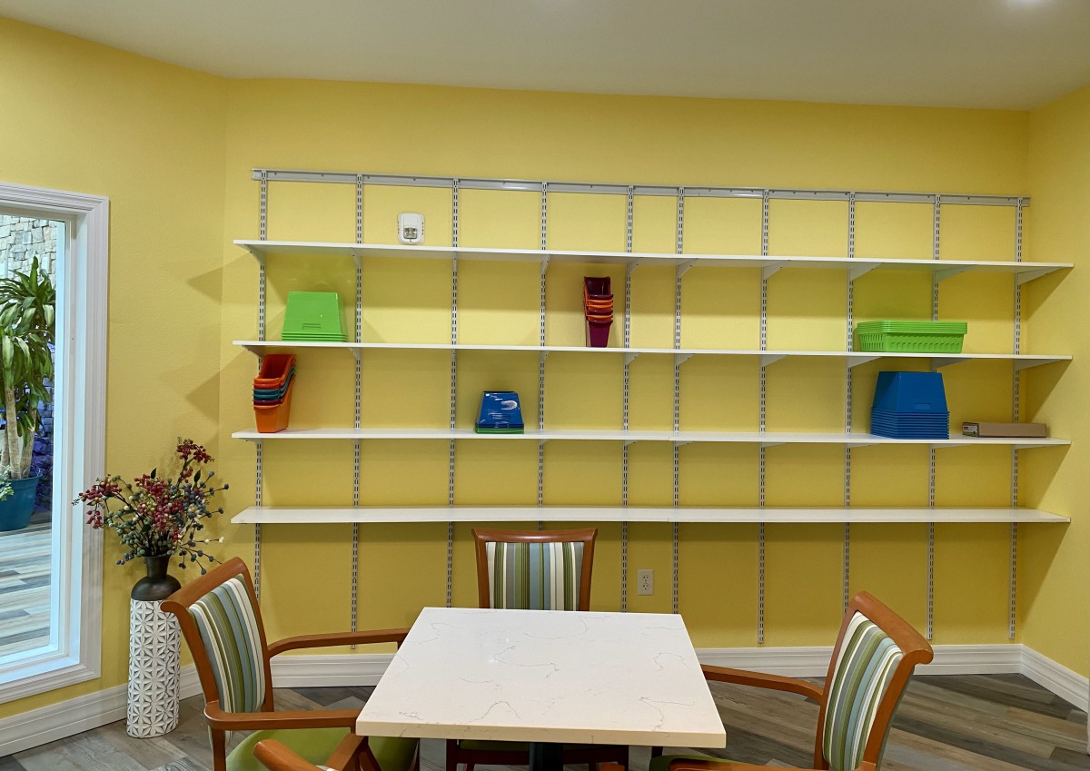 A room with a yellow wall features a large shelving unit adorned with colorful containers, accompanied by a small table and four chairs.