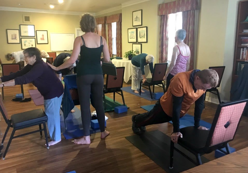 A group of adults participates in a yoga class using chairs for support.