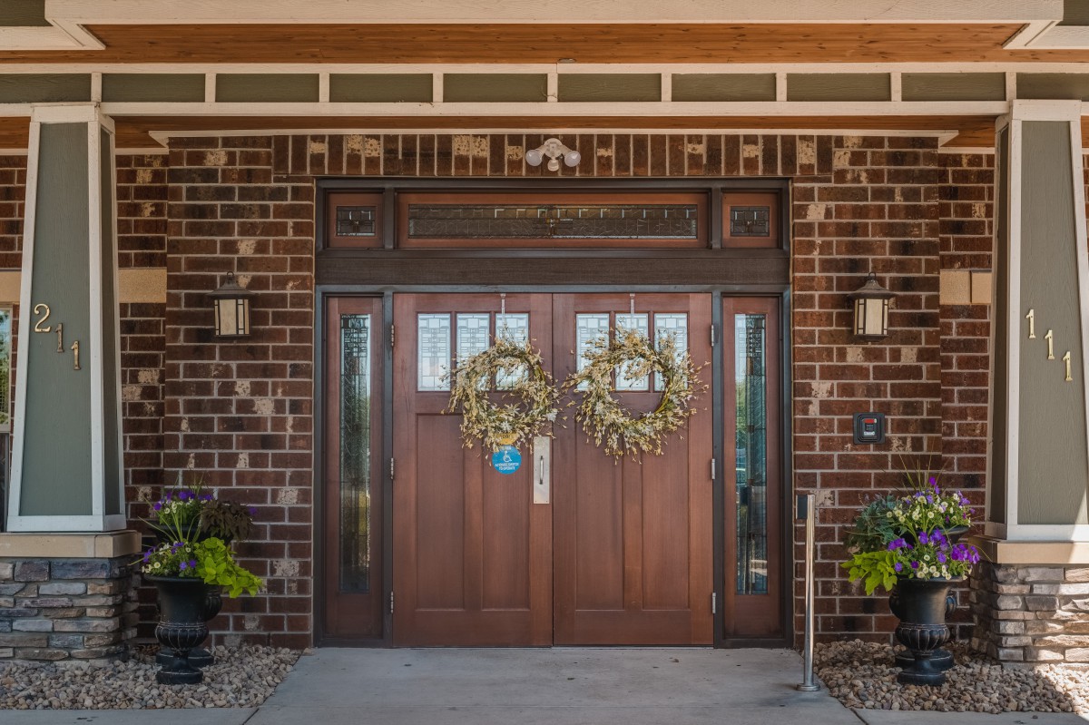 A double wooden door with decorative wreaths, set in a brick exterior with potted plants on either side.