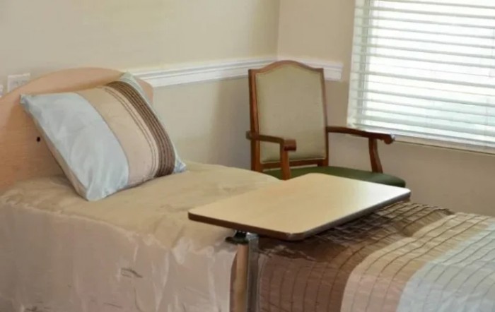 A simple, well-furnished room featuring a bed, chair, and adjustable table.