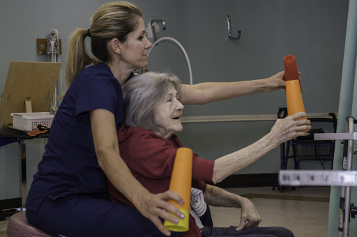 A caregiver assisting an elderly woman with a physical therapy exercise, utilizing colorful foam rollers.