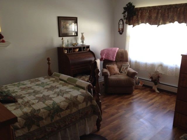 A cozy bedroom with wooden flooring, featuring a bed, recliner chair, and a desk by the window.