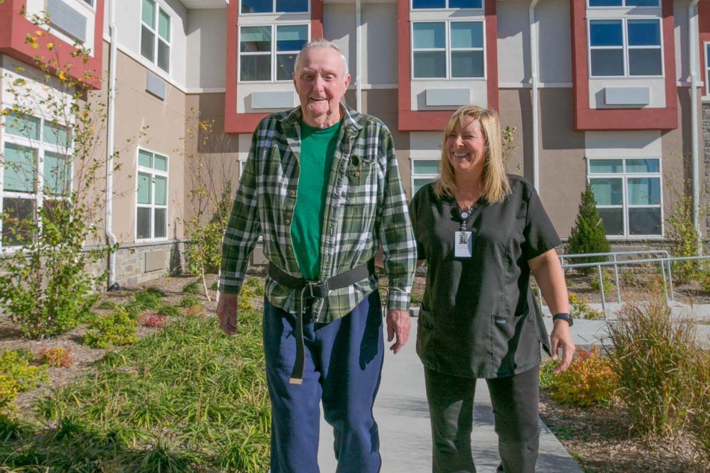 An elderly man and a woman standing outside at a care facility on a sunny day.