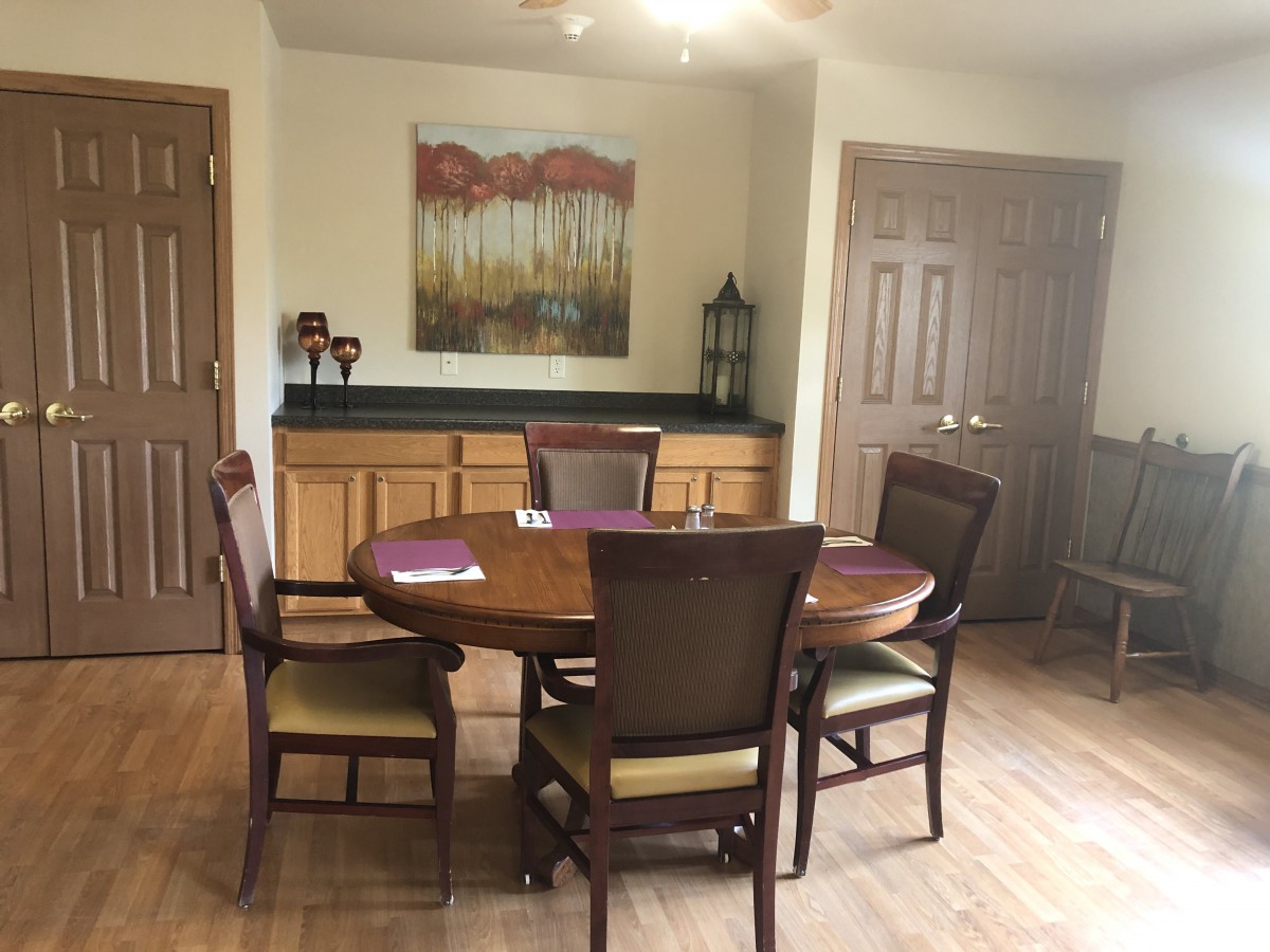 A dining room featuring a round wooden table with four chairs, a cabinet in the background, and a decorative painting on the wall.