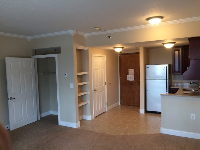 An empty apartment interior featuring a small kitchen with a fridge, cabinets, and light fixtures.