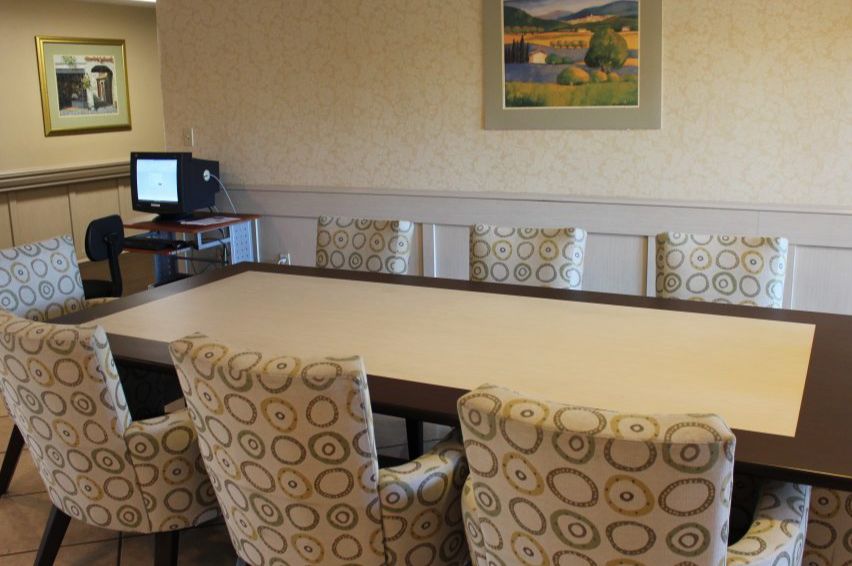A conference room with a large meeting table surrounded by patterned chairs, a computer in the corner, and a landscape painting on the wall.