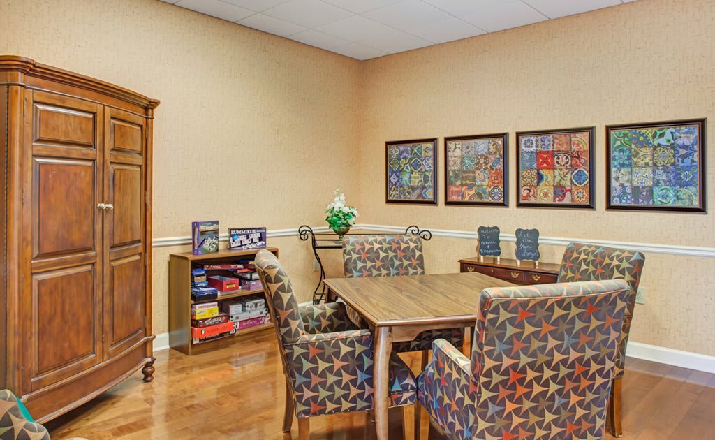 A cozy, well-decorated room featuring a wooden table with patterned chairs, artwork on the walls, and a bookshelf filled with games and books.