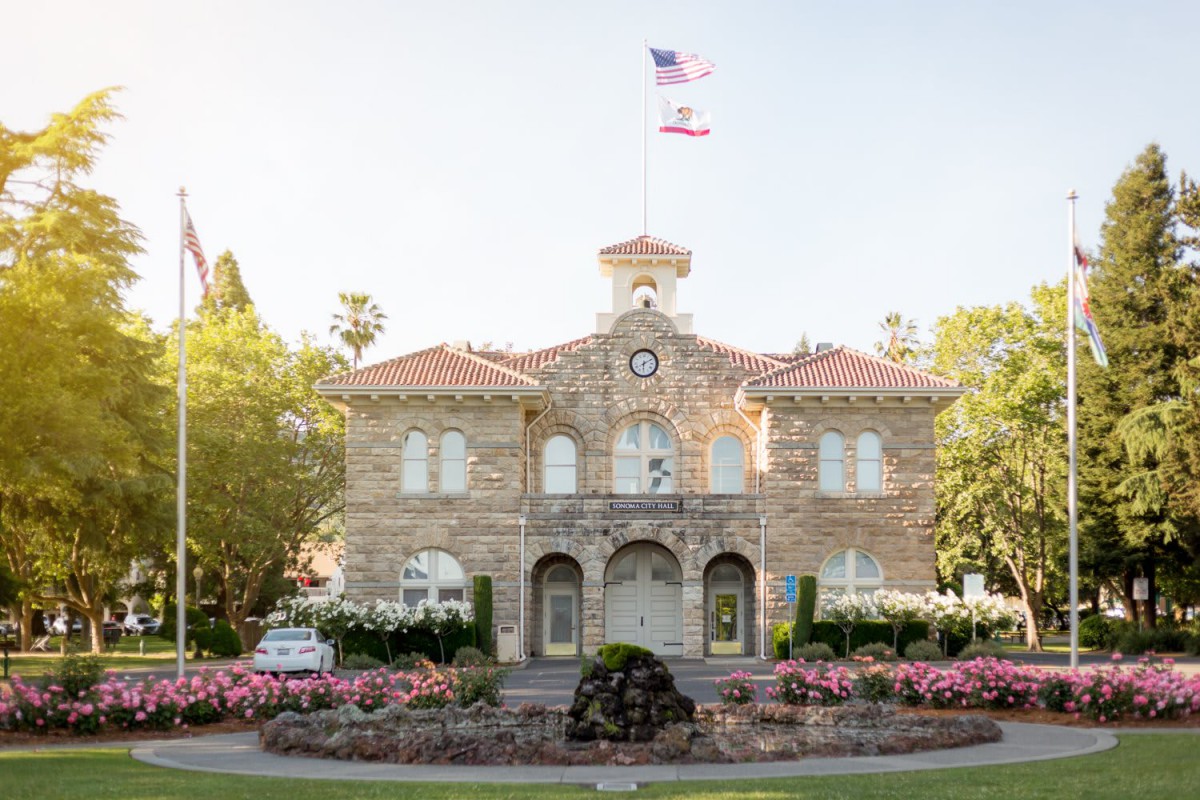 A historic stone building with an American flag flying, surrounded by a well-maintained garden on a sunny day.