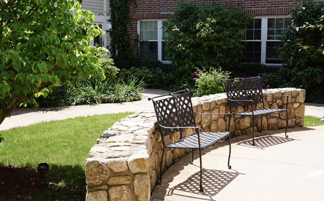 A peaceful garden area featuring metal chairs alongside a curved stone wall, surrounded by lush greenery.