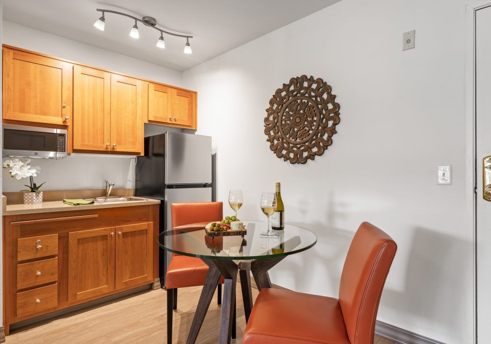 A modern kitchen and dining area with wooden cabinetry, stainless steel appliances, and a stylish glass-top table accompanied by two orange chairs.