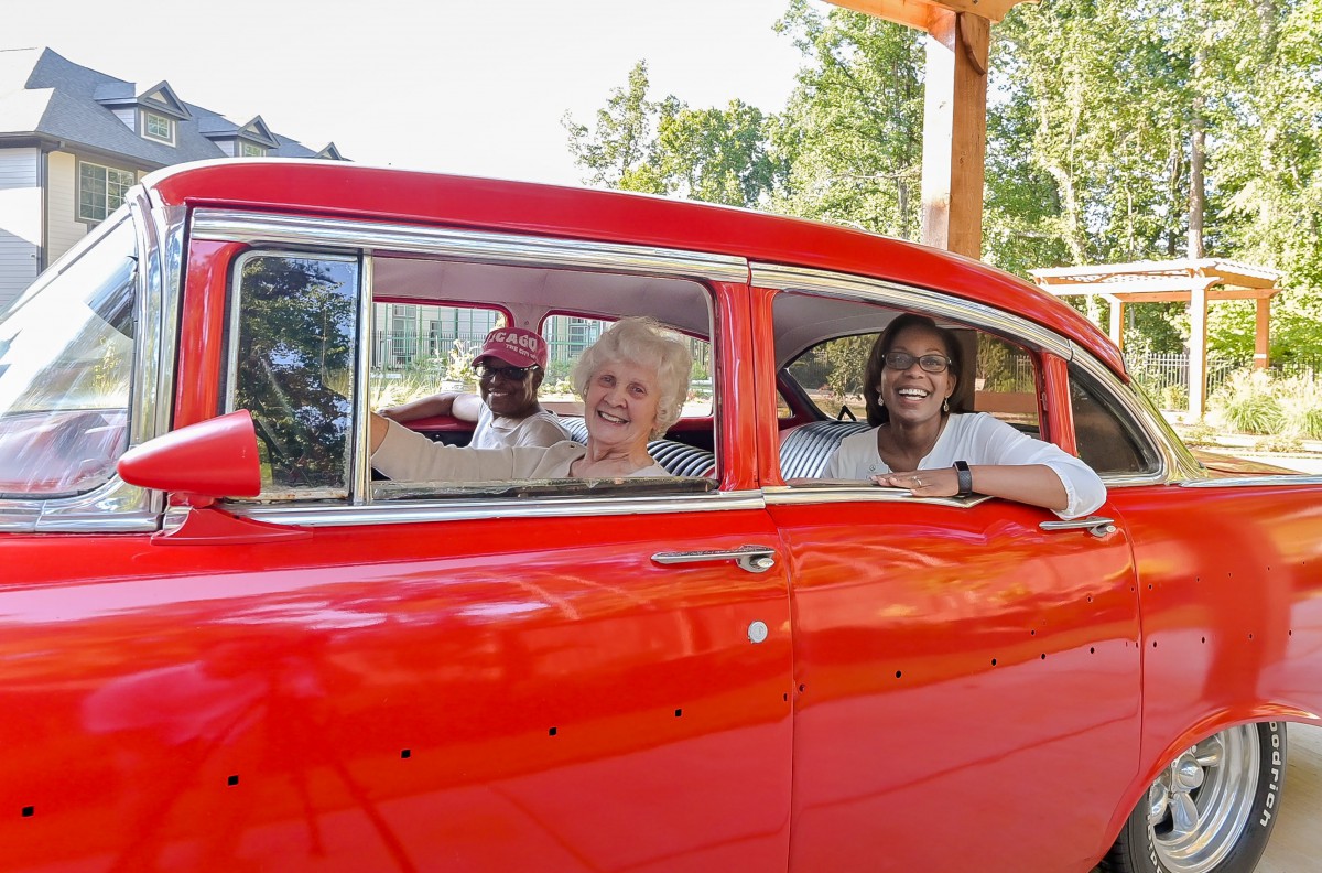 A group of people smiling while sitting inside a vintage red car outdoors.