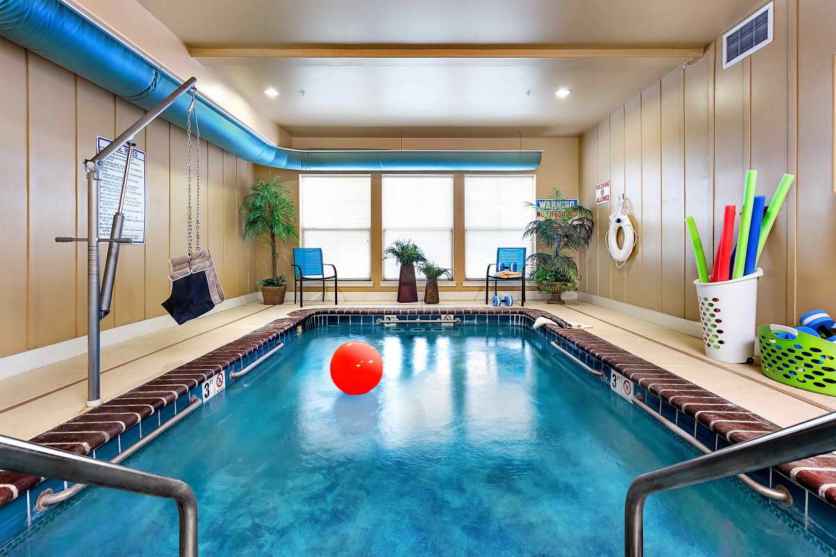 A modern indoor swimming pool with a red ball, surrounded by chairs and plants, with large windows letting in natural light.