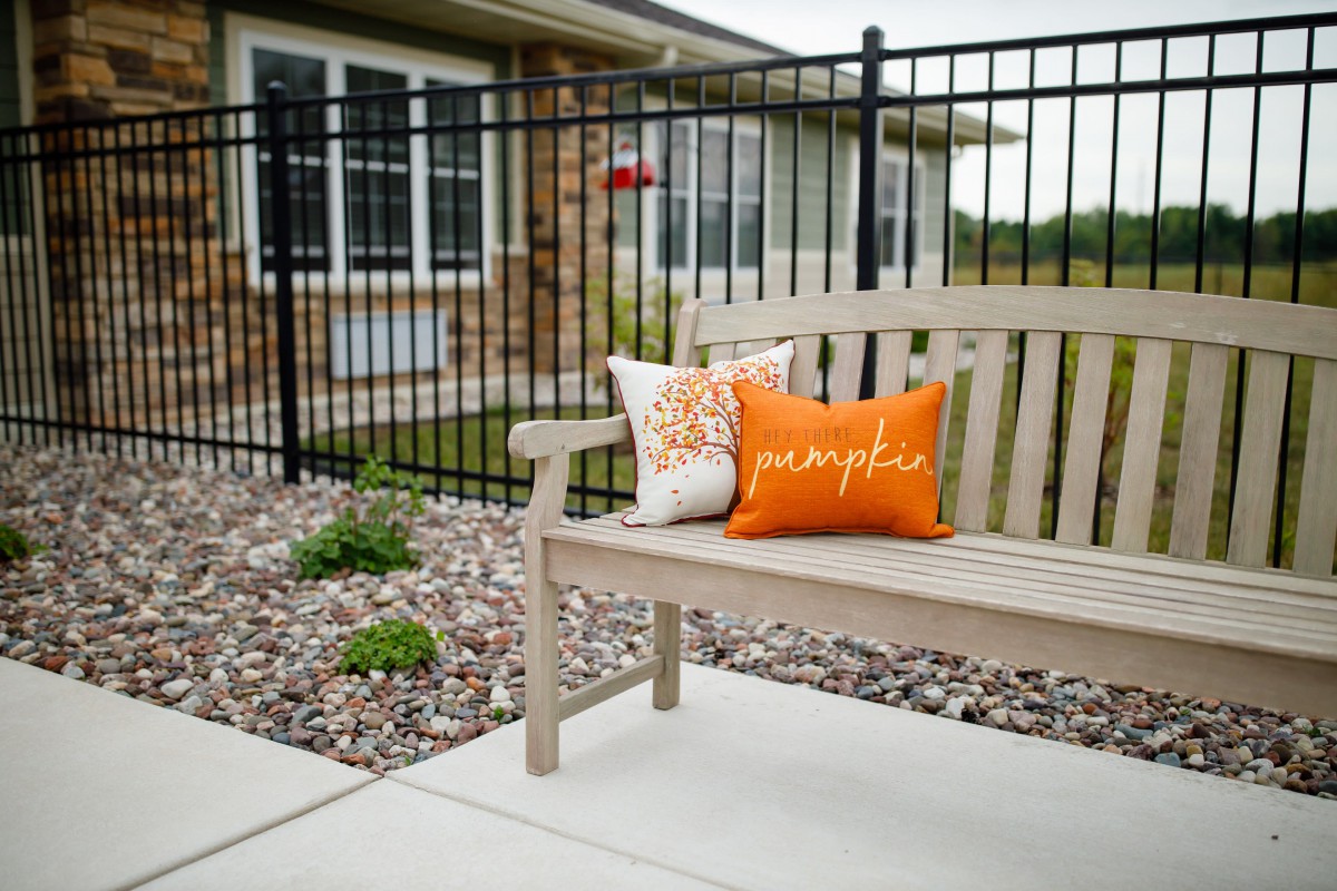 A wooden bench with decorative cushions sits on a patio, framed by a garden and fenced yard.