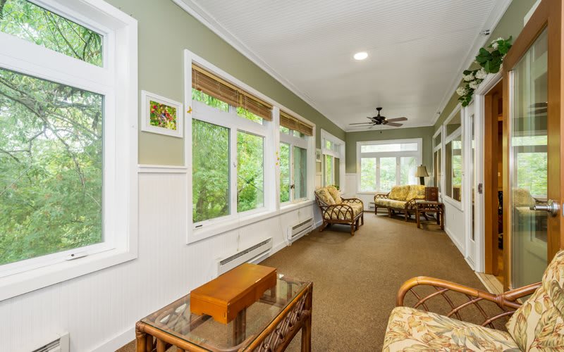 A bright sunroom with large windows, cozy furniture, and a view of the greenery outside.