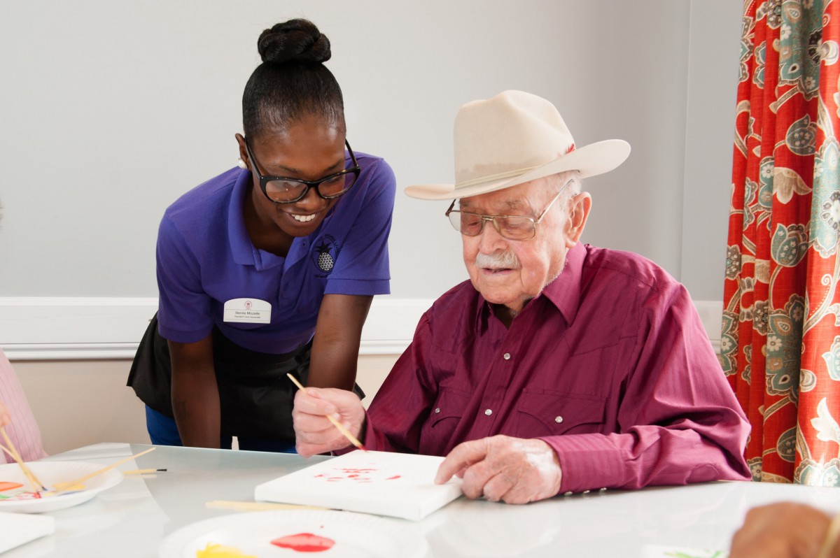 An elderly man wearing a hat is painting with the assistance of a caregiver in a nursing home.