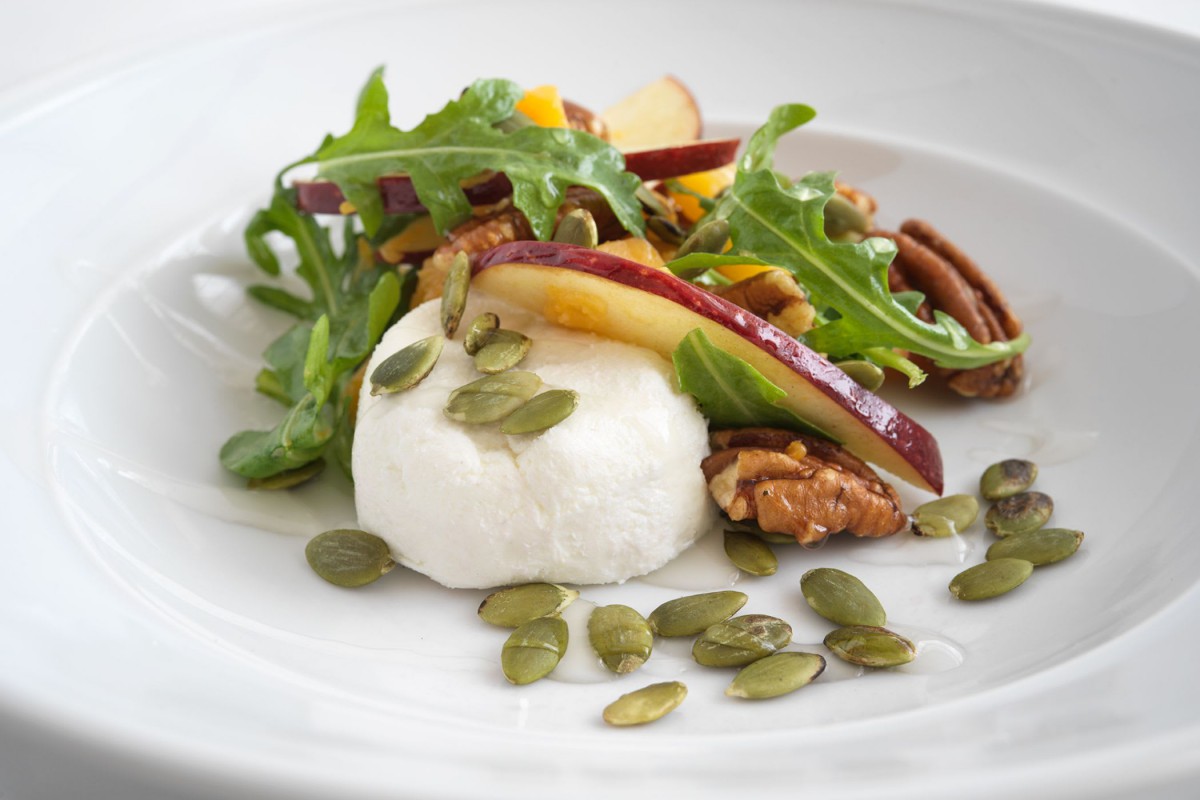 A gourmet salad featuring arugula, sliced pears, pecans, goat cheese, and pumpkin seeds, elegantly presented on a white plate.