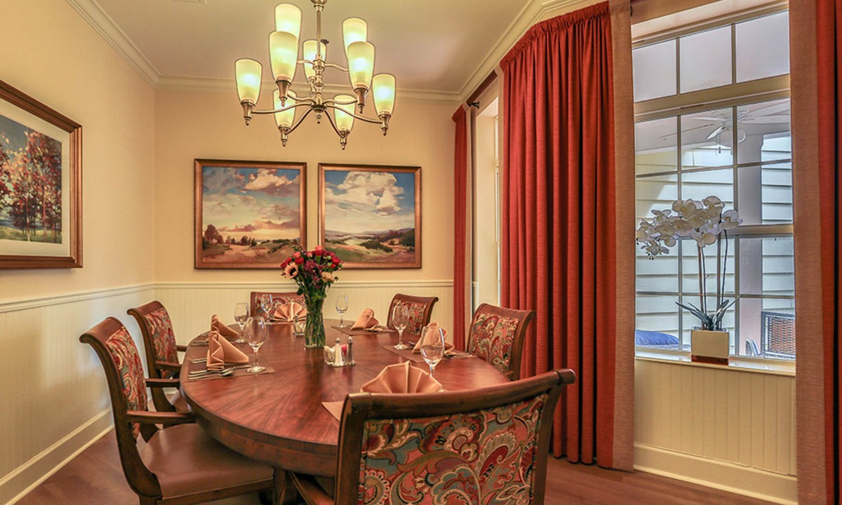 An elegant dining room with a wooden table, surrounded by chairs, and lit by a chandelier. The room features red curtains and landscape paintings on the wall.