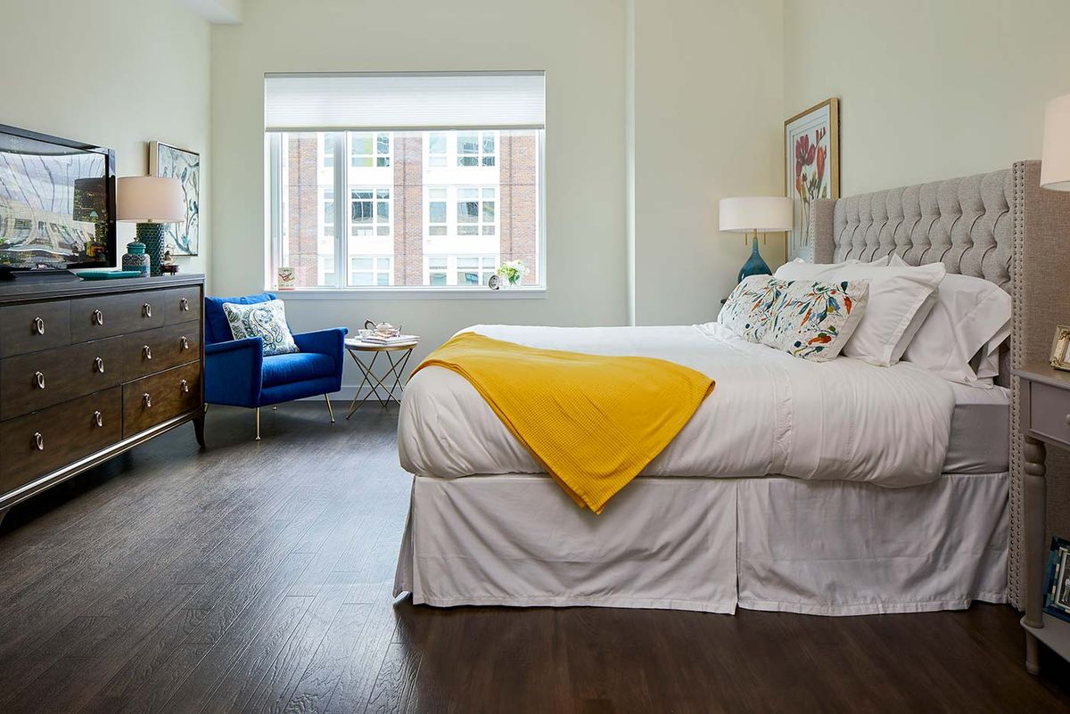 A bright and modern bedroom with a spacious bed adorned with a yellow blanket, featuring hardwood floors and an armchair by the window.