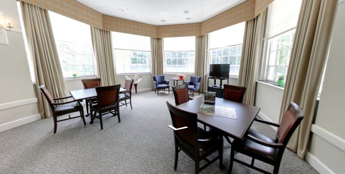 A bright dining room with multiple tables and chairs, featuring large windows with curtains.