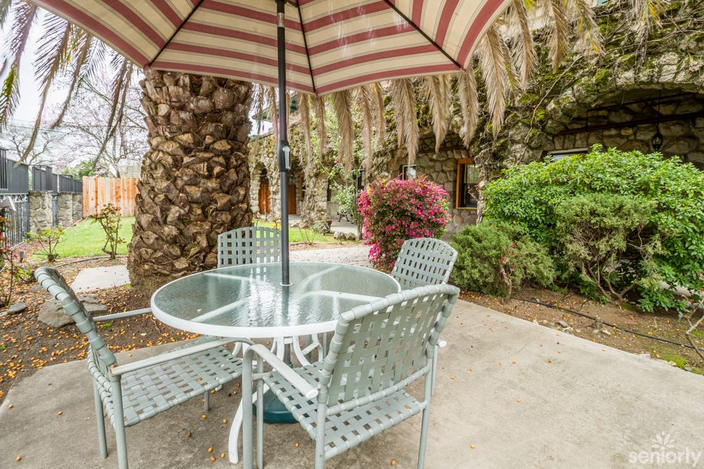 A cozy outdoor patio with a table, chairs, and an umbrella surrounded by lush greenery.