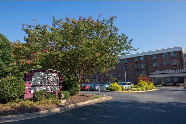 A hotel entrance with a sign, landscaped greenery, and parking area.
