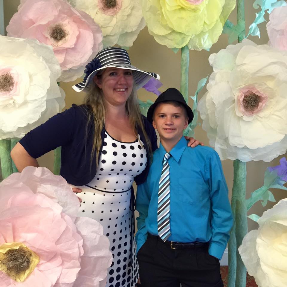 A woman and a boy in formal attire pose in front of a decorative backdrop featuring large paper flowers.
