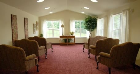 A cozy room with chairs arranged around a small table, featuring large windows and carpeted flooring.