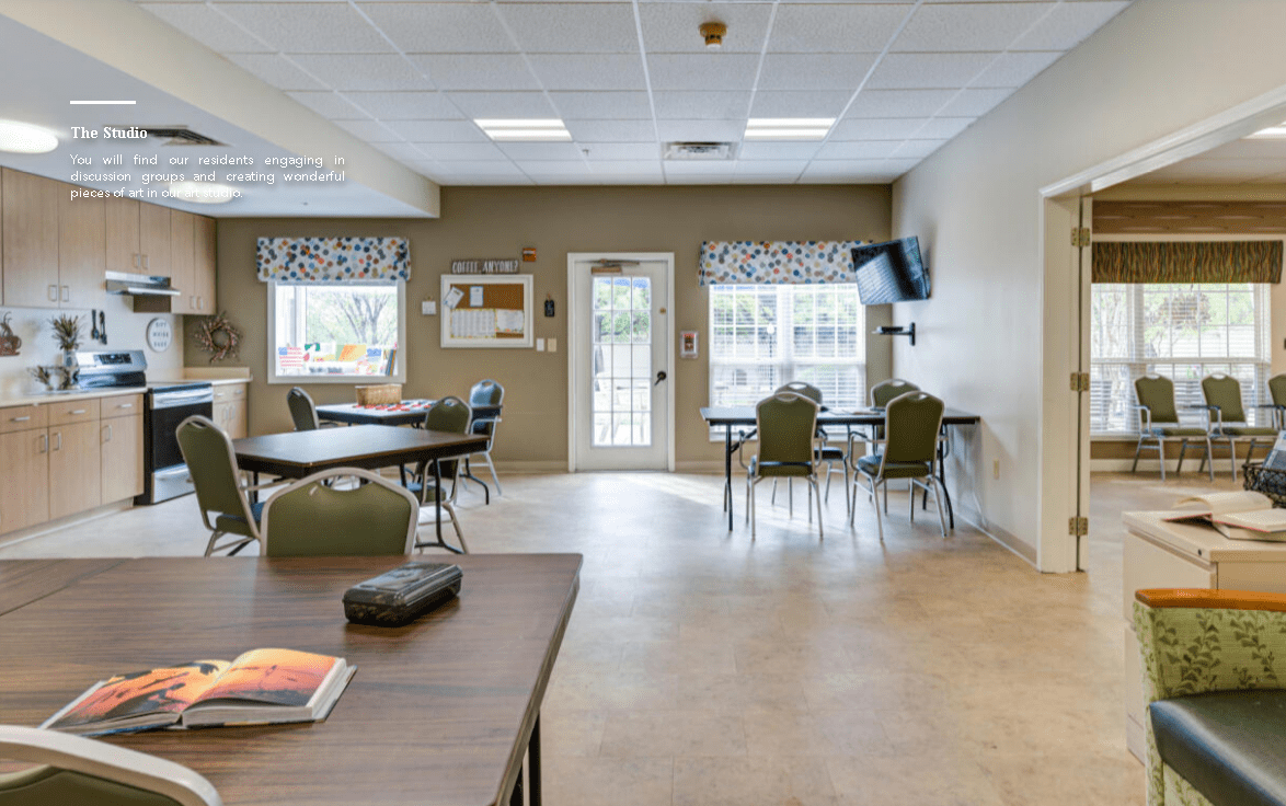 A well-lit community room featuring multiple tables and chairs, with a television mounted on the wall.