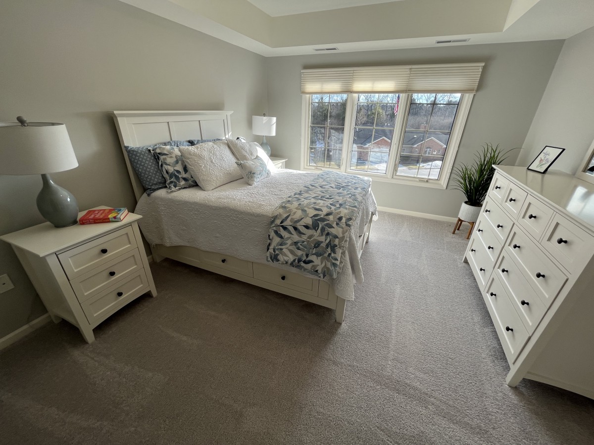 A cozy bedroom with a neatly made bed, two nightstands, and a dresser bathed in natural light from large windows.