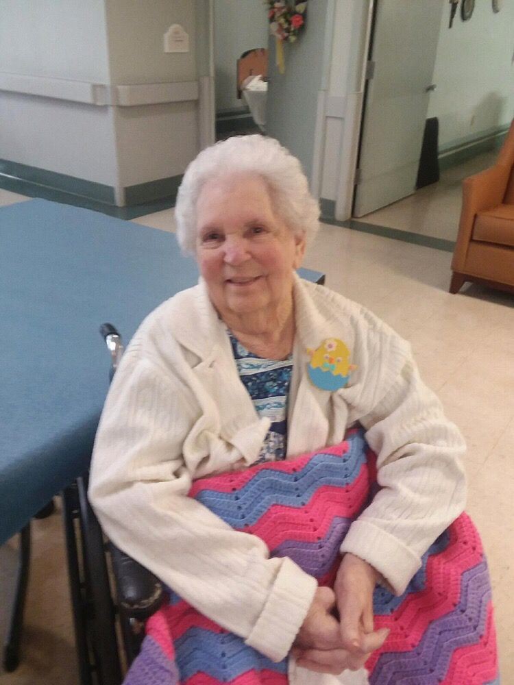An elderly woman sitting in a wheelchair, smiling and holding a colorful blanket.