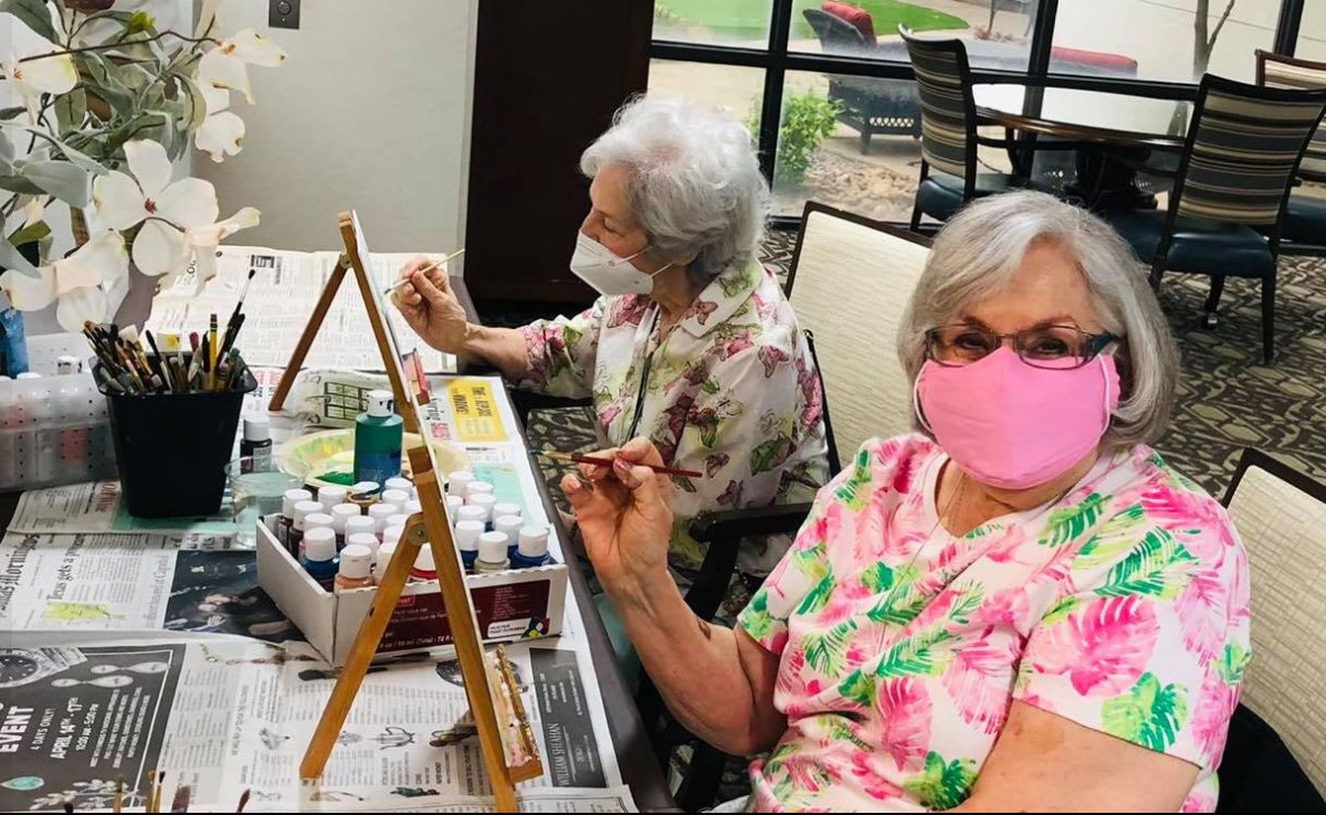 Two seniors wearing masks engage in a painting activity indoors.