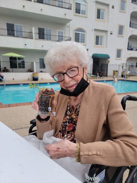 An elderly woman sits by the pool holding a dessert, with a face mask resting below her chin.