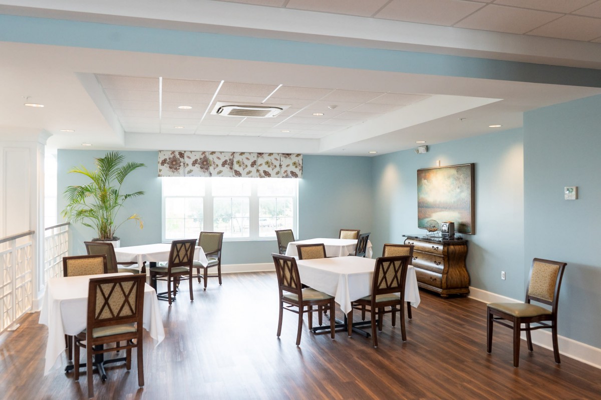 A bright and airy dining room with neatly arranged tables and chairs, featuring large windows with floral curtains and a wooden floor.