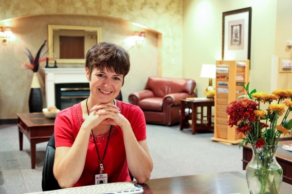 A woman in a red shirt is sitting at a desk in an office area, smiling at the camera.