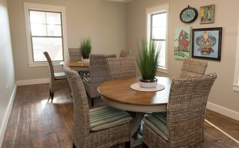 A cozy dining area featuring round wooden tables, wicker chairs, and potted plants, bathed in natural light from nearby windows.