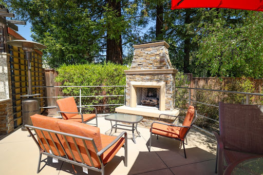 A cozy outdoor patio featuring comfortable seating and a charming stone fireplace, surrounded by lush greenery.