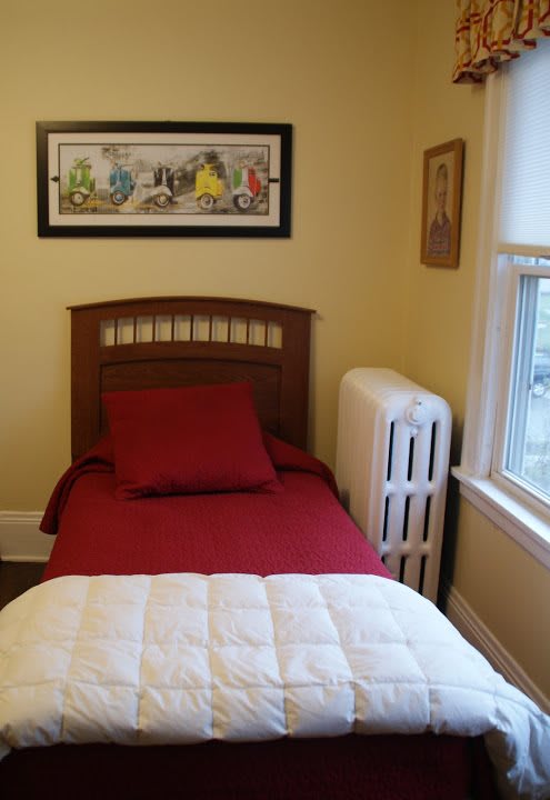 A cozy bedroom featuring a single bed with red bedding, a radiator by the window, and wall art.